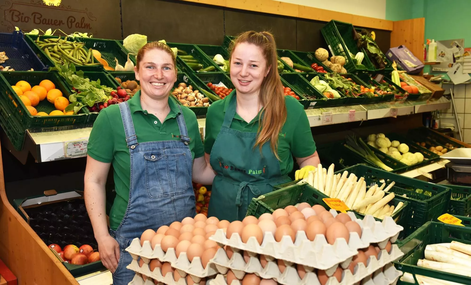 Zwei Mitarbeiterinnen im Bio-Hofladen Palm vor Regalen mit frischem Bio-Obst, Gemüse, Eiern und Spargel.